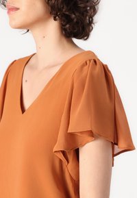 Femme portant un chemisier couleur rouille avec des manches papillon et un col en V, vue de profil sur un fond blanc uni.