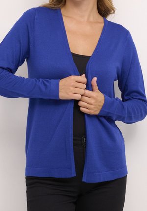 Femme portant un cardigan bleu par-dessus un haut noir et un pantalon noir, fermant le cardigan avec ses mains.
