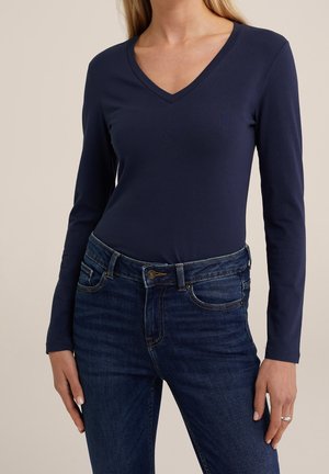 Vrouw draagt een marineblauw, lang mouwig V-hals shirt, ingestopt in donkerblauwe spijkerbroek, staand tegen een effen beige achtergrond.