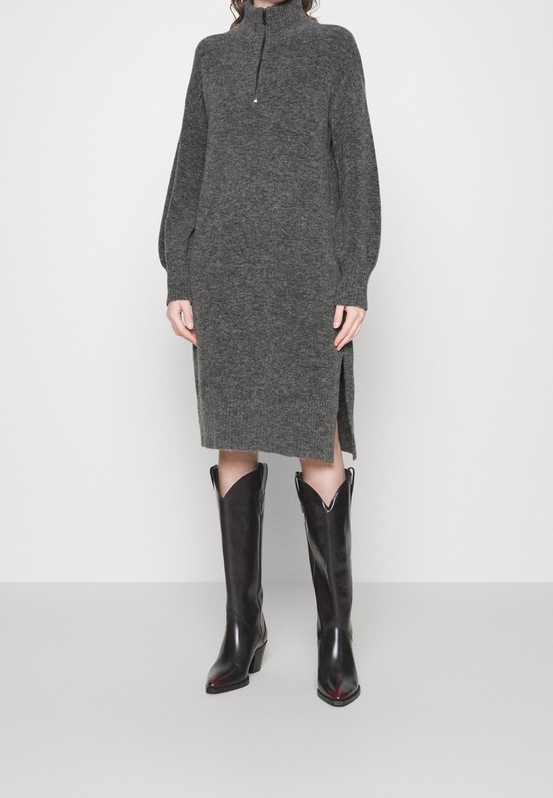 Femme portant une robe en maille grise longueur genou avec une fermeture éclair au quart et une fente latérale, associée à des bottes en cuir noires montant jusqu'aux genoux.