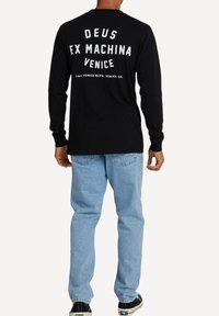 Schwarzes Langarm-T-Shirt mit weißen Text auf der Rückseite, in lockerer Passform, kombiniert mit hellblauen Jeans und schwarzen Sneakers.