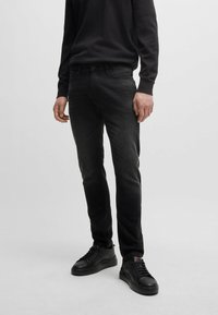 Black jeans in a slim fit with a faded design, paired with black sneakers. The fabric has a smooth texture and minimal visible stitching.