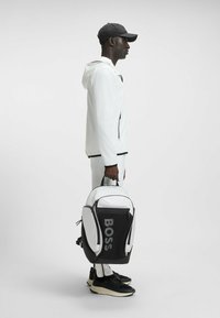 Vit sportjacka med svarta accenter, svart keps och en svart-vit ryggsäck med "BOSS"-logotyp, i kombination med svarta sneakers.