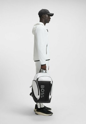 Witte sportjack met zwarte accenten, zwarte pet en een zwart-wit rugzak met "BOSS" logo, gecombineerd met zwarte sneakers.