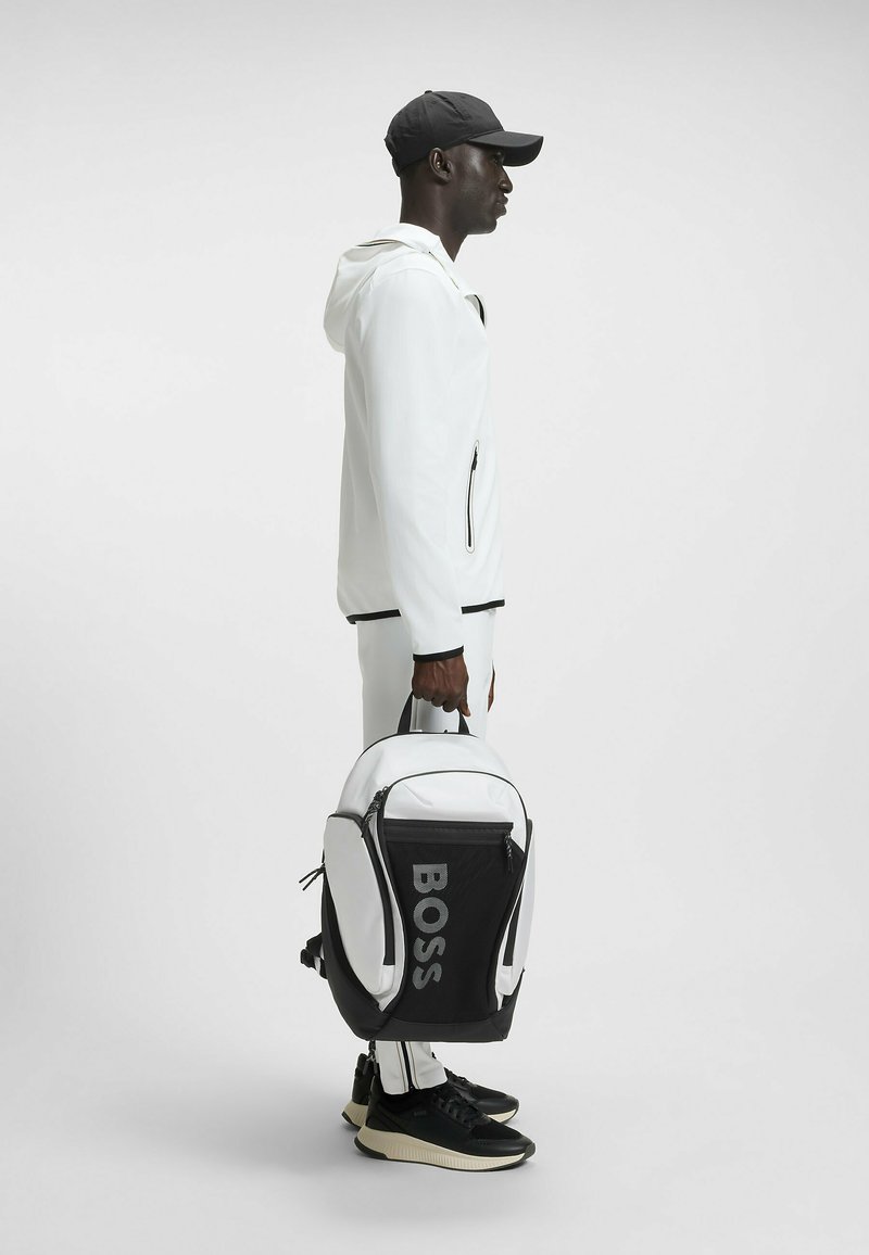 Vit sportjacka med svarta accenter, svart keps och en svart-vit ryggsäck med "BOSS"-logotyp, i kombination med svarta sneakers.