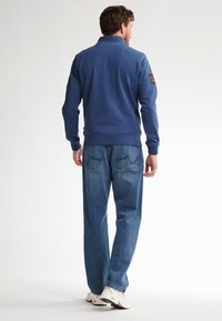 Felpa blu con collo alto e maniche lunghe, abbinata a jeans azzurri larghi e sneakers bianche. Punti spalla visibili.
