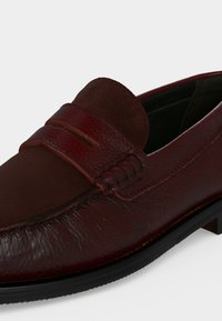 Mocassin en cuir brun foncé avec détail de bride penny et semelle en caoutchouc noir, vu en plan rapproché de côté.