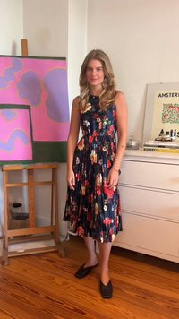 Rochie florală, plisată, în nuanța navy, cu modele colorate de flori. Este purtată cu pantofi negri cu bot pătrat. Fundalul include artă abstractă.