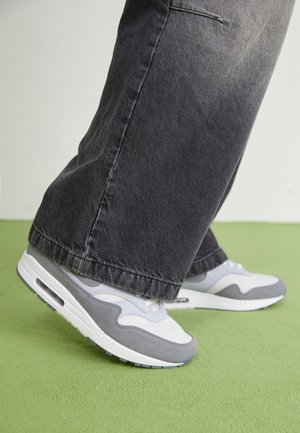 Osoba nosząca szaro-białe trampki oraz czarne jeansy z szerokimi nogawkami stoi na zielonym dywanie.