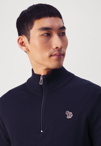 Tengerészkék, cipzáros nyakú pulóver puha kötött anyagból, kontrasztos, hímzett zebra logóval a bal mellen és bordás gallérral a fokozott textúra érdekében.