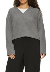 Grauer V-Ausschnitt-Pullover mit langen Ärmeln, aus weichem Stoff gefertigt, mit geripptem Halsauschnitt und lässiger Passform, kombiniert mit schwarzen Hosen.