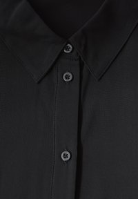 Camicia nera con colletto e tre bottoni neri visibili lungo la patta frontale, cuciture dettagliate sul colletto e sull'area dei bottoni.