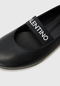 Sapatilha plana de couro preto com bico arredondado, apresentando uma tira elástica preta com o texto "VALENTINO" em branco. Textura suave e design minimalista.