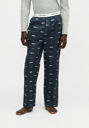 Homme portant un pantalon de pyjama Calvin Klein bleu marine avec des logos blancs et un t-shirt gris à manches longues, debout pieds nus sur fond blanc.