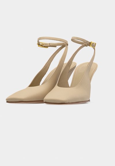 Μπεζ γόβες slingback με μυτερή μύτη, λεπτά λουράκια στον αστράγαλο και χρυσαφένιες αγκράφες σε λευκό φόντο.