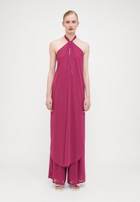Robe longue à dos nu en chiffon rose vif. Présente un détail en forme de clé et un design superposé. Tissu fluide créant de douces drapés.