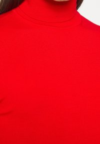 Roter gerippter Rollkragenpullover, der ein enganliegendes Design mit glatter Textur und hohem Kragen zeigt. Der Stoff scheint weich und dehnbar zu sein.