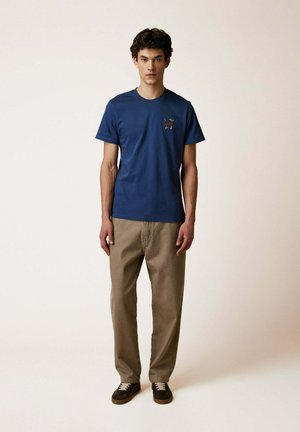 Giovane uomo in piedi contro uno sfondo semplice, indossa una maglietta blu con una piccola grafica sul petto, pantaloni beige e scarpe da ginnastica nere.