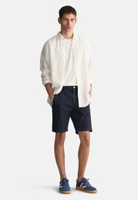 Camisa blanca de lino de manga larga, camiseta blanca debajo, pantalones cortos azul marino, zapatillas de ante azules con suelas marrones. Ajuste casual, sin patrones visibles.