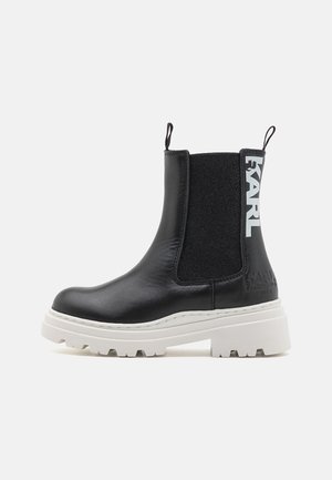 Bottines Chelsea en cuir noir avec des panneaux élastiques sur les côtés et une semelle épaisse blanche. Présente un logo embossé et des tirettes pour un enfilage facile.