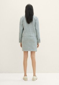 Blazer in tweed azzurro chiaro con gonna coordinata. Presenta un design aderente, una texture sottile e bottoni decorativi sulle maniche. Scarpe casual visibili.