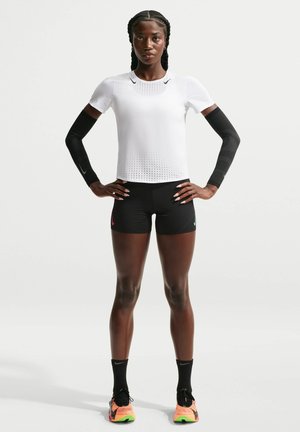 Femme athlétique debout, les mains sur les hanches, portant un haut blanc à manches courtes, un short noir, des manchons pour les bras, des chaussettes noires et des chaussures de course orange.