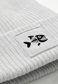 Light gray ribbed knit beanie featuring a rectangular label with a black fish skeleton graphic. Soft texture, form-fitting design.