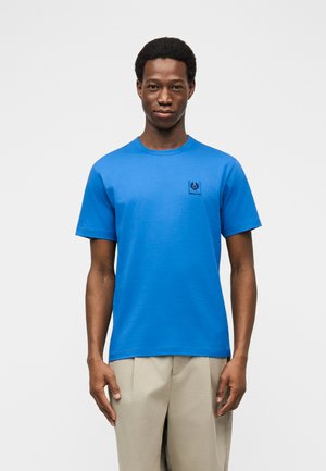 Muž nosící jasně modré tričko s krátkým rukávem a malým černým logem na levé straně hrudi, béžové kalhoty na jednobarevném bílém pozadí.