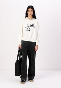 Felpa bianca con il testo "Saddle Up" in nero, abbinata a pantaloni larghi grigio scuro. Il modello tiene una borsa tote nera, indossando scarpe nere.