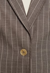 Blazer marron à fines rayures beige verticales avec un seul grand bouton beige moucheté pour fermer le devant.