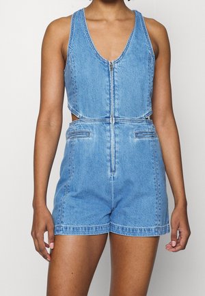Woman wearing sleeveless blue denim romper with front zipper and cutout sides, standing against a plain light background.