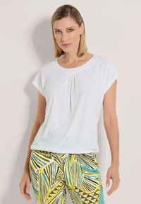 Weiße Bluse mit kurzen Ärmeln, Rundhalsausschnitt und Faltenarchitektur an der Vorderseite. Kombiniert mit einer gemusterten Hose in Grün und Blau mit Blättermotiven.