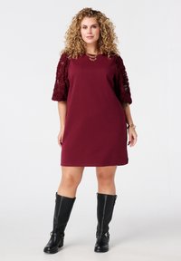Robe bordeaux avec des manches florales texturées, longueur genoux, assortie à des bottes noires jusqu'aux genoux. Un collier en or souligne le décolleté.
