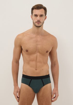 Homme musclé aux cheveux bruns courts portant un slip turquoise avec une ceinture noire, debout devant un fond clair uni.