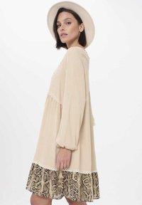 Crème-kleurige jurk met uitlopende mouwen, kantaccenten en een slangenprint volantszoom. Lichtgewicht stof, A-lijn vorm.