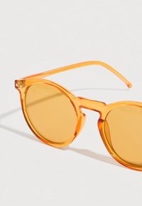 Στρογγυλά γυαλιά ηλίου με πορτοκαλί διαφανή πλαίσια και καφέ φακούς. Διαθέτουν χοντρή γέφυρα και λεπτά μπράτσα.