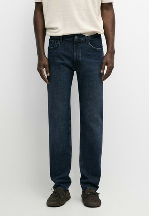 Mand iført mørkeblå straight-leg jeans, beige kortærmet skjorte og mørkebrune snørestøvler, der står mod en ensfarvet baggrund.