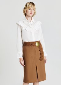 Blusa bianca con colletto a volant, abbinata a una gonna in suede marrone con intagli circolari e una grande fibbia decorativa.