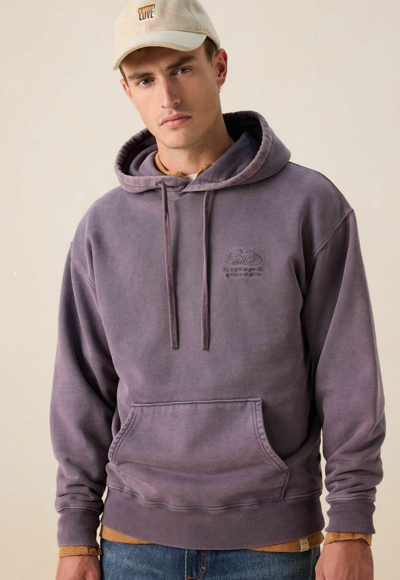 Een jonge man in een vervaagde paarse hoodie met een voorzak en een beige pet met de borduursel "LOVE", staand tegen een effen lichte achtergrond.