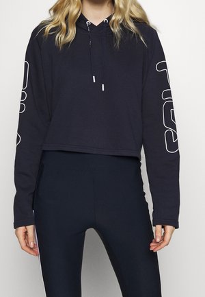 Femme portant un sweat à capuche raccourci noir avec des lignes graphiques blanches sur les manches et un legging noir ajusté, debout devant un fond uni.