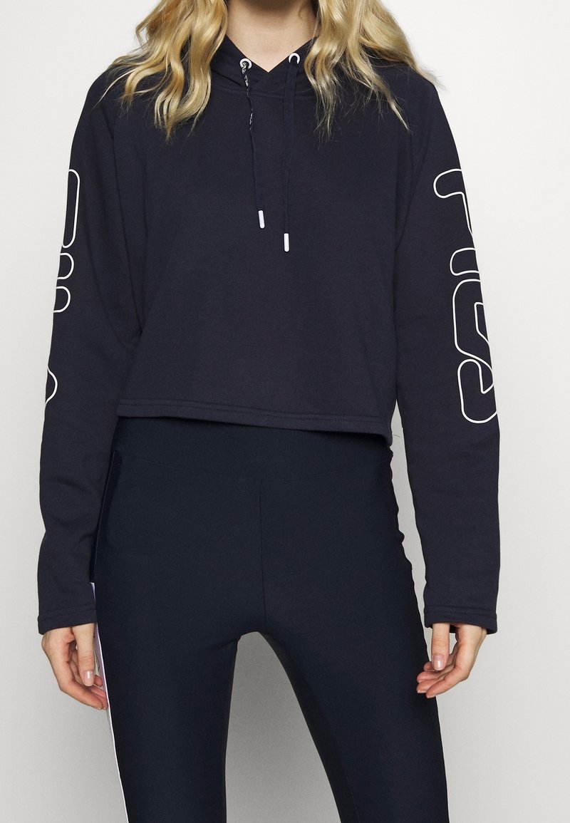 Femme portant un sweat à capuche raccourci noir avec des lignes graphiques blanches sur les manches et un legging noir ajusté, debout devant un fond uni.