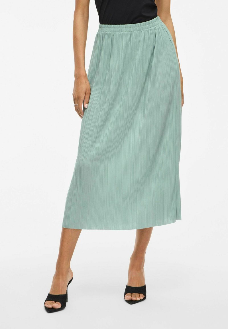 Minttu vihreä, taitettu midi-hame elastisella vyötäröllä ja sileällä pinnalla, yhdistettynä mustiin avonaisiin korkokenkiin.