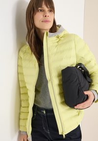 Doudoune jaune clair avec capuche, motif matelassé et fermeture éclair argentée. Pull beige en dessous. Sac noir tenu dans une main.