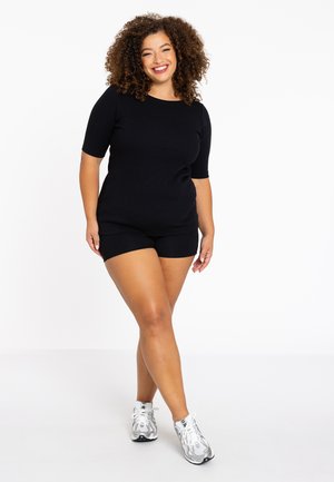 Sort kortærmet top og matchende shorts, lavet af et blødt stof. Kombineret med grå og hvide sneakers. Modellen har krøllet hår.