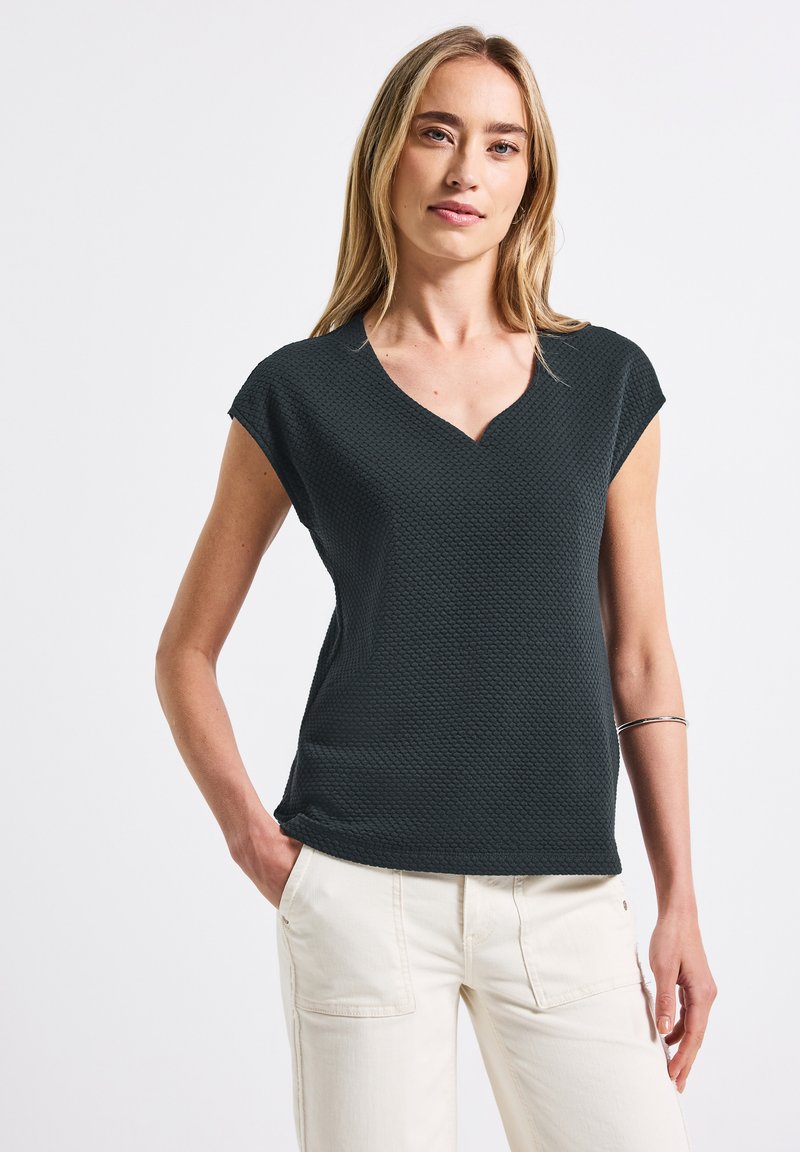 Mörkgrön kortärmad topp med en texturerad bikupamönster och V-ringning, kombinerad med ljusa byxor.