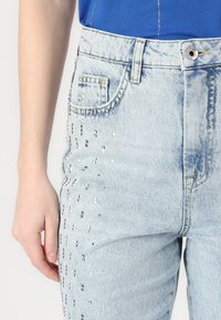 Vaqueros de denim azul claro con una cintura alta, que presentan lentejuelas plateadas decorativas dispuestas en líneas verticales en la parte delantera.
