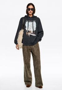 Ciemnoszara bluza z kapturem z grafiką przedstawiającą krajobraz z drzewami, zestawiona z oliwkowozielonymi szerokimi spodniami w wzory i brązowymi zamszowymi butami.