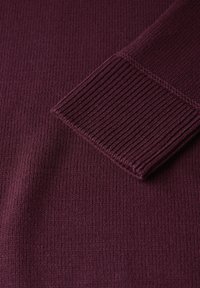 Pull en maille bordeaux avec une texture lisse, présentant des poignets et un ourlet côtelés. Le gros plan met en valeur les belles coutures et la qualité du tissu.