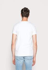 Weißes, kurzärmeliges T-Shirt mit Rundhalsausschnitt, aus Baumwolle, von hinten betrachtet. Enganliegendes Design, glatte Textur. Getragen mit hellblauen Jeans.