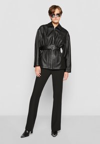 Giubbotto in finta pelle nera con vita regolabile, chiusura con zip e borchie decorative, abbinato a pantaloni neri aderenti e stivali neri a punta.